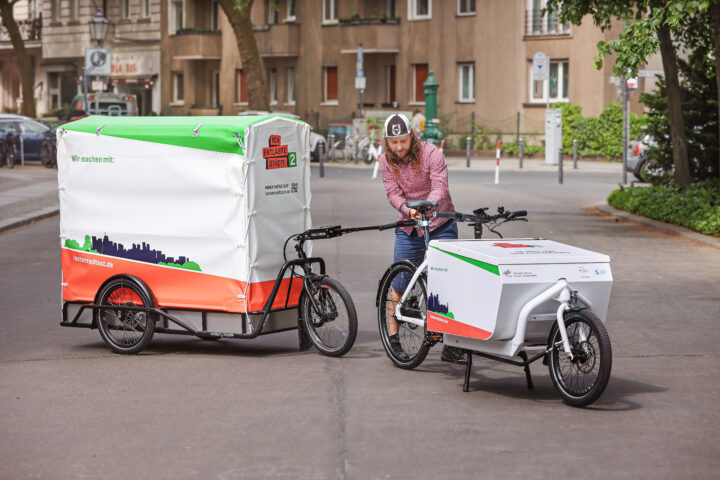 An das Lastenrad Larry vs Harry wird ein dreirädriger Fahrradanhänger der Firma Carla Cargo angespannt. Der Anhänger hat einen hohen Aufbau mit einer Plane.