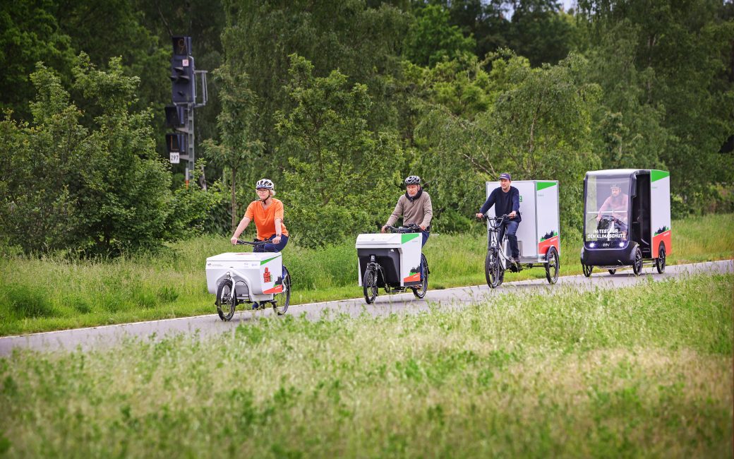 Auf einem Radweg fahren hintereinander vier Lastenräder: Mit Transportkoffer vorne oder hinten, mit zwei, drei oder vier Rädern