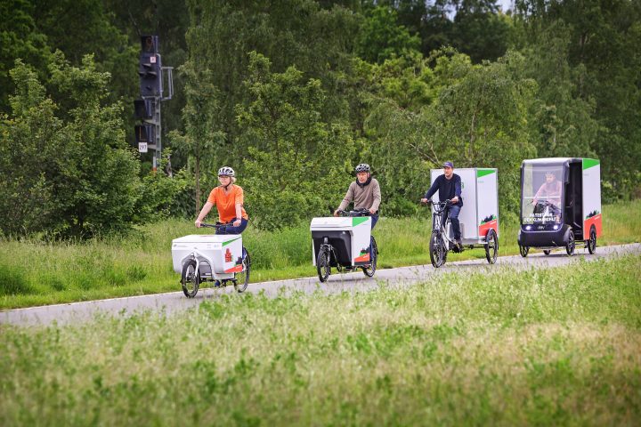 Auf einem Radweg fahren hintereinander vier Lastenräder: Mit Transportkoffer vorne oder hinten, mit zwei, drei oder vier Rädern