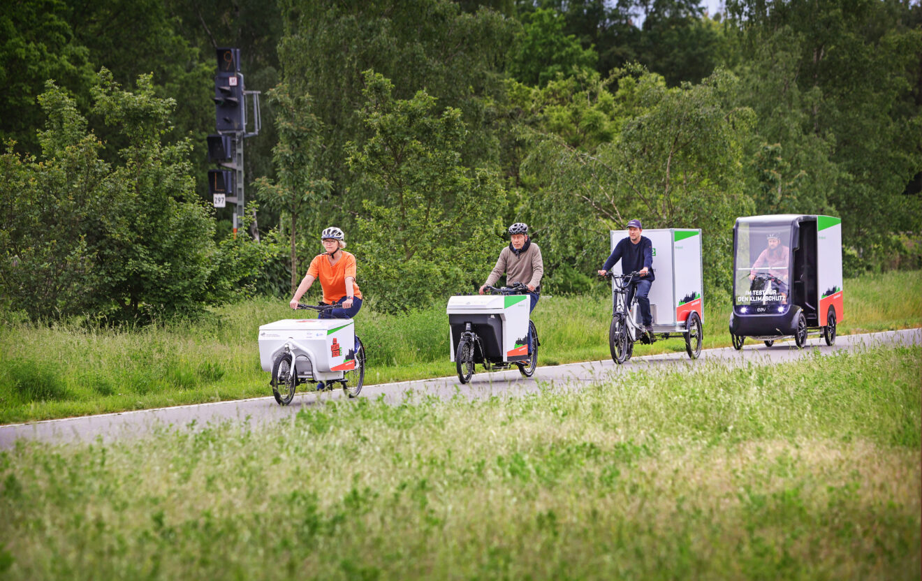 Auf einem Radweg fahren hintereinander vier Lastenräder: Mit Transportkoffer vorne oder hinten, mit zwei, drei oder vier Rädern